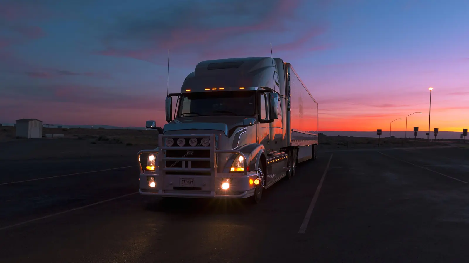 A tractor-trailer with a sleeper cab and its headlights on, parked at a rest area at dusk under an orange and purple sky